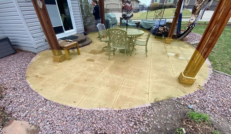 A completed concrete patio with tile surface installed under a pergola structure, surrounded by gravel landscaping and backed by residential structures.