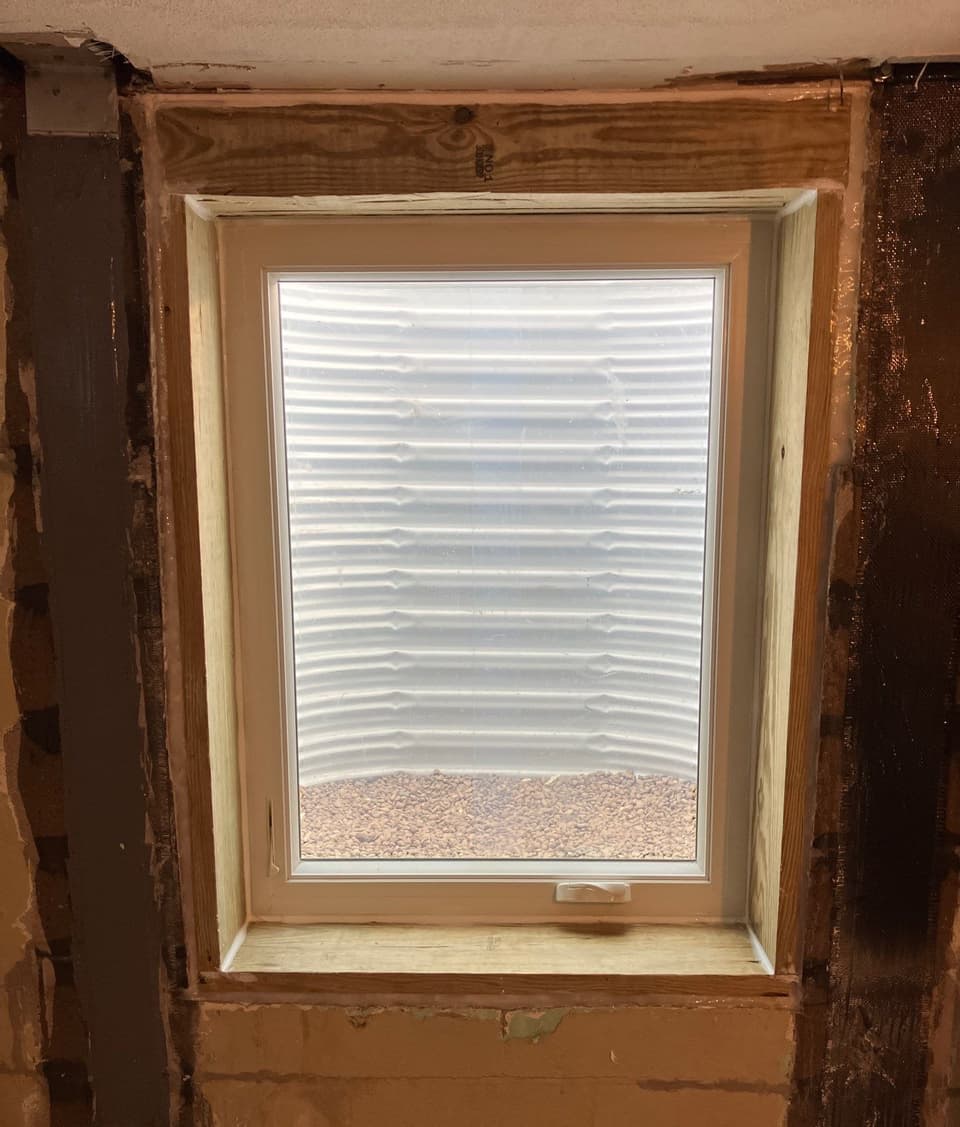 A newly installed egress window with light-colored wood frame and sill viewing out to a beach/sandy area with ocean waves in the background.