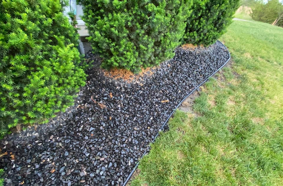 A foundation drainage system with black landscape fabric edging separating a gravel drainage bed from the surrounding lawn adjacent to evergreen shrubs.