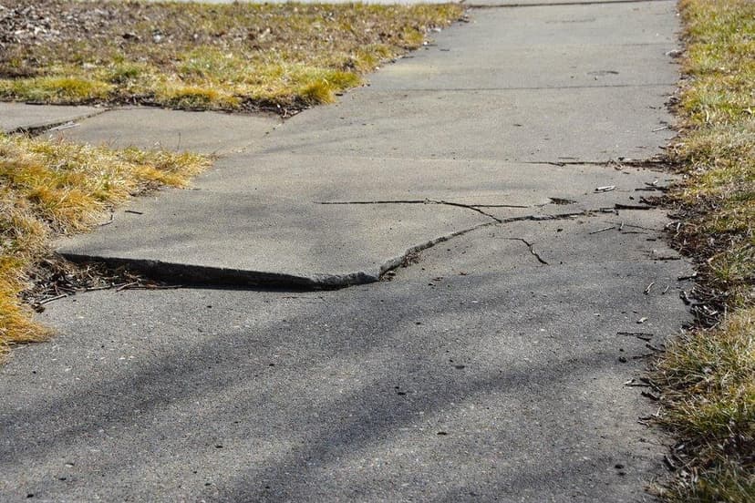 Cracked and uneven concrete sidewalk with signs of soil settlement.