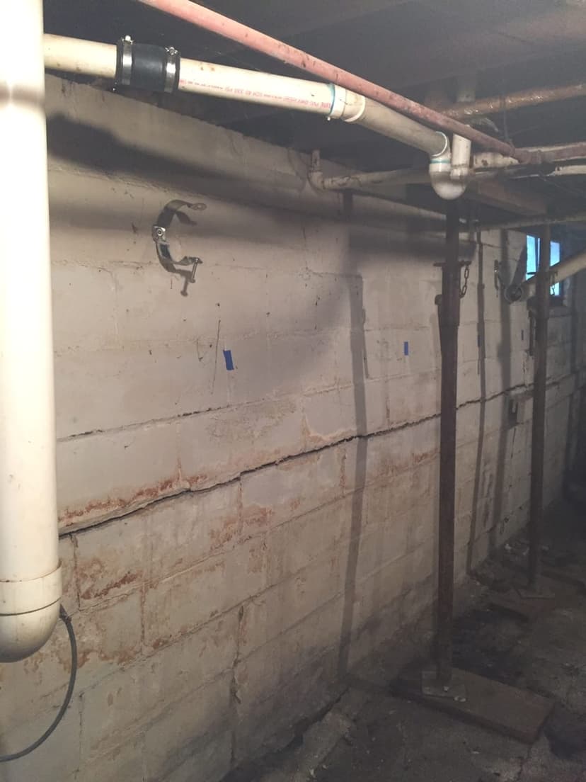 Real photo of a basement block wall with a horizontal crack line marked with small blue painter's tape flags, overhead PVC and copper plumbing pipes visible, despite the filename the wall bowing itsel